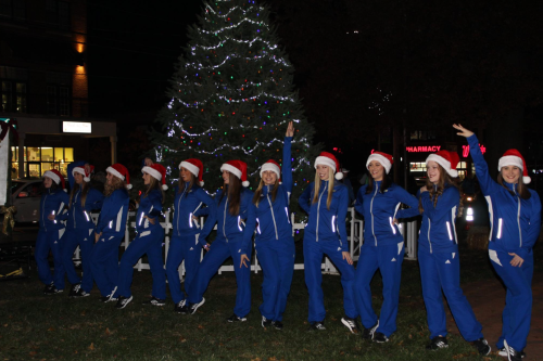 A group of people in blue outfits and Santa hats poses joyfully in front of a decorated Christmas tree at night.