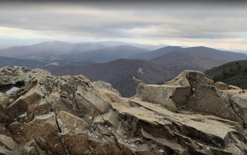 A rocky mountain summit with a panoramic view of rolling hills and valleys under a cloudy sky.