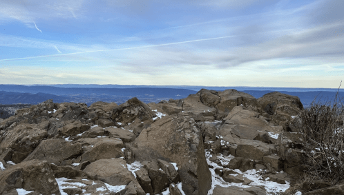 Rocky mountain summit with scattered snow, overlooking a vast landscape under a cloudy sky.