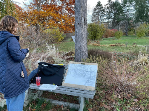 A person stands by a wooden table in a garden, sketching with a drink beside them, surrounded by autumn foliage.
