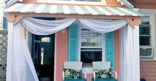 A charming porch with white drapes, teal shutters, and two floral-patterned chairs, creating a cozy outdoor space.