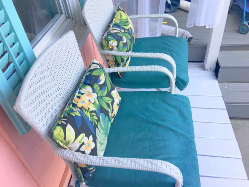 Two white wicker chairs with green cushions and floral pillows, placed on a wooden porch with pastel-colored walls.