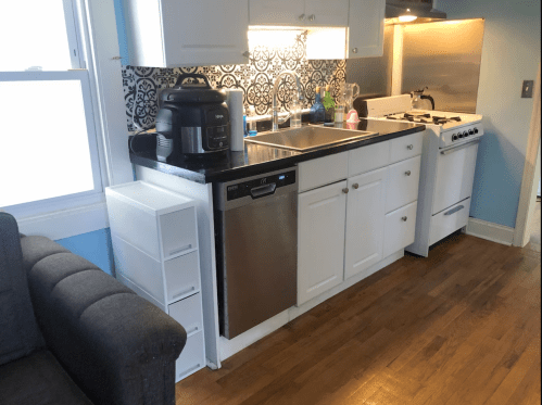 A kitchen with a sink, dishwasher, stove, and an air fryer on the counter, next to a gray couch and storage drawers.