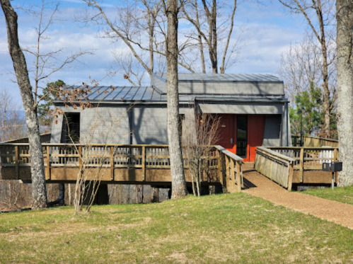 A modern house with a wooden deck, surrounded by trees and grass, under a blue sky with scattered clouds.