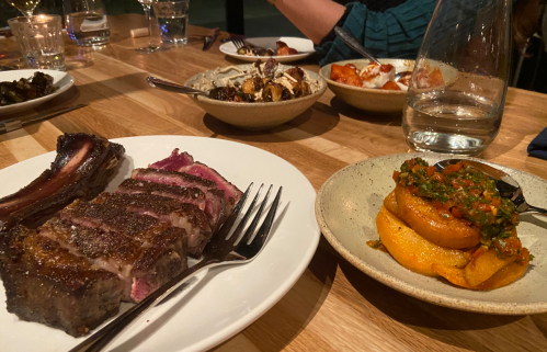 A plate with sliced steak and a side of roasted squash topped with salsa, surrounded by other dishes on a wooden table.