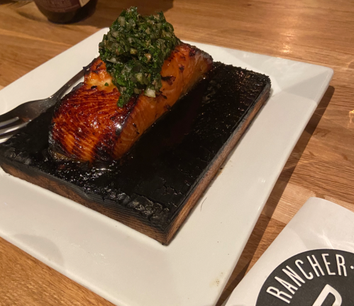 Grilled salmon fillet topped with green herb sauce, served on a charred wooden plank on a white plate.