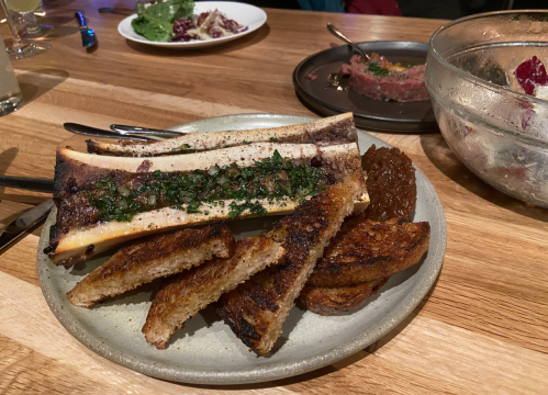 A plate featuring grilled bone marrow topped with herbs, accompanied by toasted bread and a side of meat spread.
