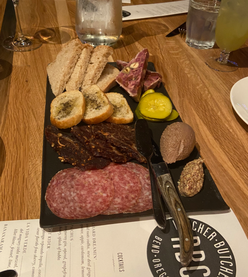 A charcuterie board featuring meats, spreads, pickles, and bread on a wooden table.