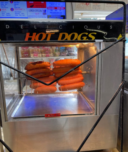 A hot dog warmer displaying several hot dogs on skewers, with a menu board visible in the background.