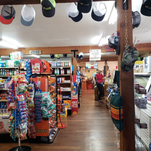 Interior of a convenience store with shelves of snacks, drinks, and hats hanging from the ceiling. A customer is browsing.