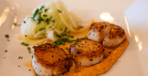 Three perfectly seared scallops on a bed of sauce, accompanied by a side of cucumber noodles and garnished with herbs.