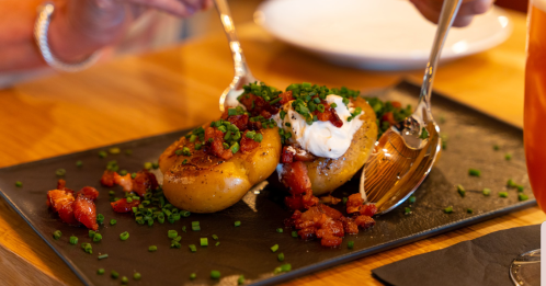 A close-up of two baked potatoes topped with bacon, sour cream, and chives on a black plate, with utensils nearby.