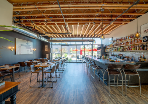 Modern restaurant interior with a bar, wooden floors, and large windows overlooking an outdoor seating area.