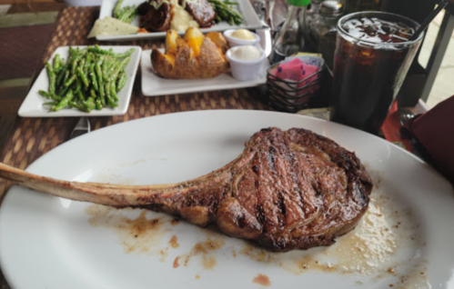 A large, grilled tomahawk steak on a white plate, with sides of green beans, baked potato, and a drink in the background.