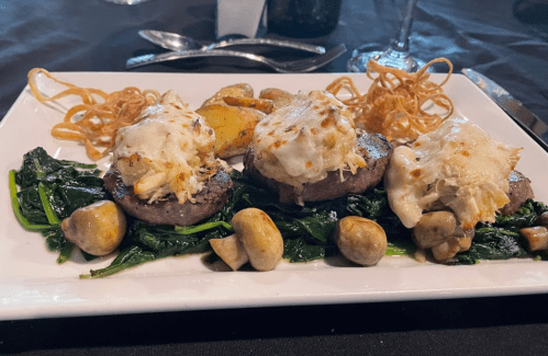 A gourmet plate featuring three beef medallions topped with crab, served on sautéed greens and roasted potatoes.