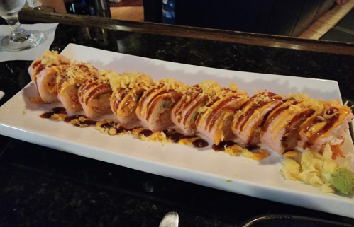 A long sushi roll topped with drizzles of sauce and crunchy bits, served on a white plate with ginger on the side.