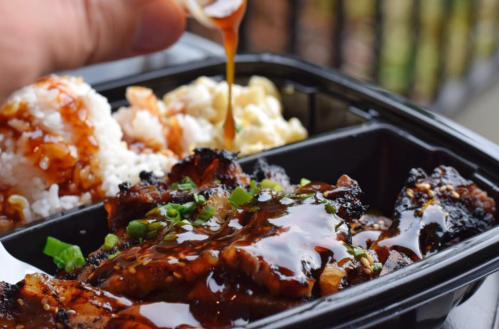 A hand drizzles sauce over a black takeout container filled with grilled meat, rice, and macaroni salad.