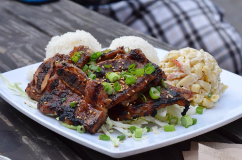 A plate of grilled ribs topped with green onions, served with rice and macaroni salad on a wooden table.