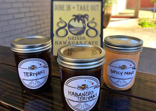 Three jars of sauces labeled Teriyaki, Habanero Teriyaki, and Spicy Mayo in front of a Hawaiian café sign.