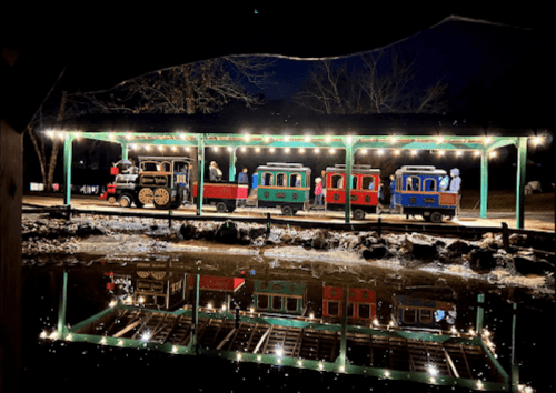 A colorful train on a platform at night, illuminated by string lights, reflecting in the water below.
