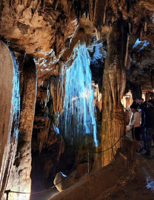 A cave interior with stalactites and a waterfall illuminated by blue light, with visitors observing the scene.