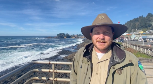 A park ranger stands by the ocean, smiling with a coastal town in the background and waves crashing against the rocks.
