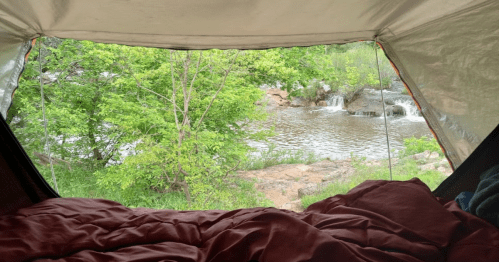 View from inside a tent, showcasing a river and lush greenery outside, with a cozy sleeping area in the foreground.
