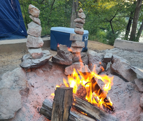 A campfire surrounded by stacked stones, with a cooler in the background and trees nearby.