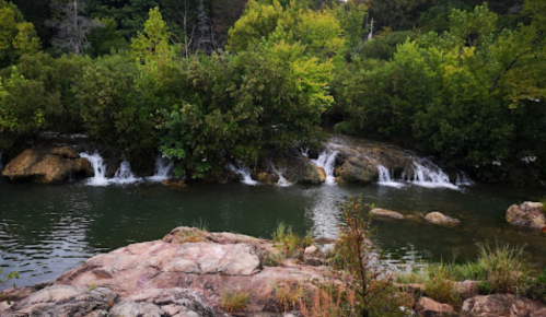 A serene landscape featuring a small waterfall cascading into a calm green pool, surrounded by lush trees and rocks.