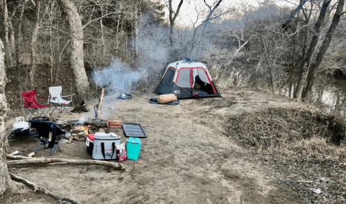 A campsite by a river with a tent, chairs, a campfire, and cooking equipment surrounded by bare trees.