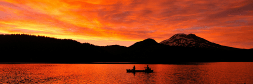 A serene sunset over a lake, with silhouettes of two people in a canoe and a snow-capped mountain in the background.