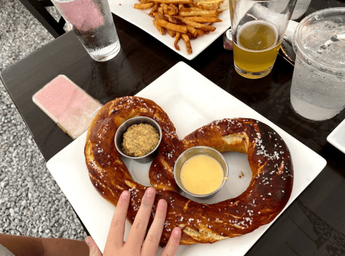 A large, twisted pretzel on a plate with two dipping sauces, accompanied by fries and drinks in the background.