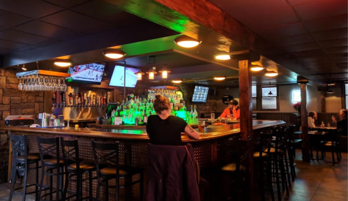 A cozy bar with a wooden counter, illuminated bottles, and patrons enjoying drinks in a warm, inviting atmosphere.