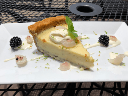 A slice of cheesecake on a white plate, garnished with mint, blackberries, and a drizzle of cream.