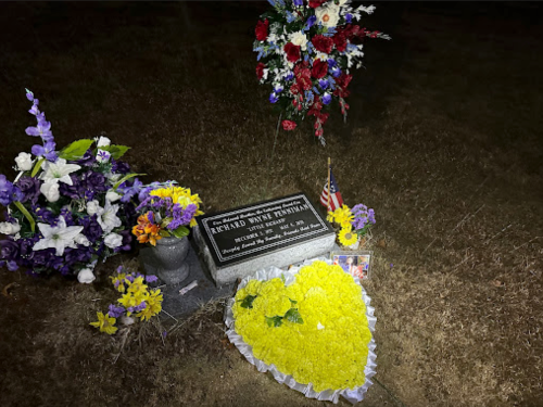 A grave adorned with flowers, a heart-shaped floral arrangement, and a small American flag.