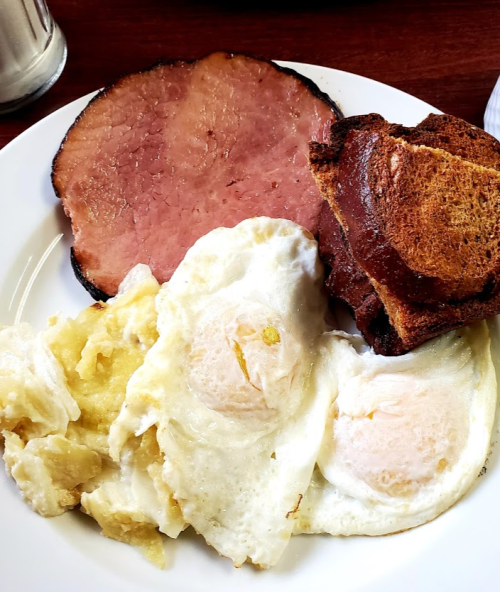 A plate with two fried eggs, a slice of ham, and a piece of toasted bread.