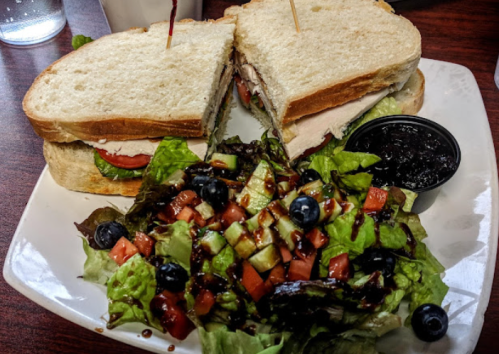 A sandwich with turkey, lettuce, and tomato, served with a side salad of mixed greens, tomatoes, cucumbers, and blueberries.