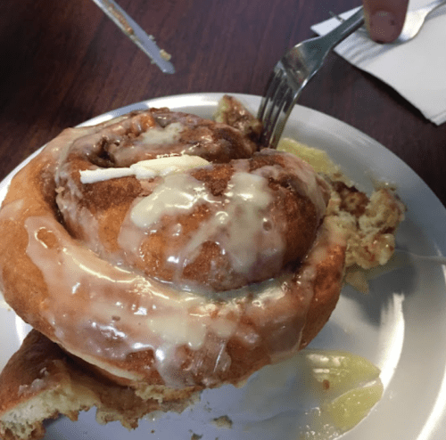 A close-up of a cinnamon roll topped with icing and a pat of butter on a white plate, with a fork beside it.