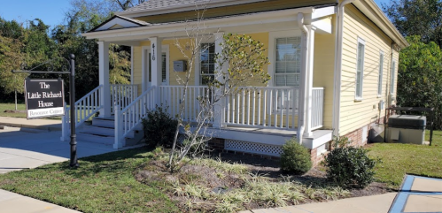 A yellow house with a porch and sign reading "The Little Richard House Resource Center" surrounded by greenery.