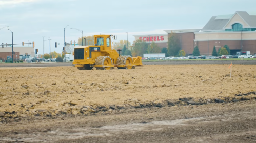 A yellow bulldozer works on a large, cleared dirt lot near a Scheels store in the background.