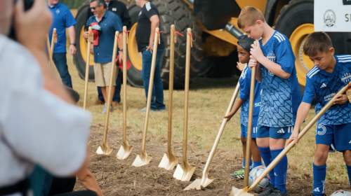 Children in blue soccer uniforms stand with shovels at a groundbreaking ceremony, surrounded by onlookers and machinery.