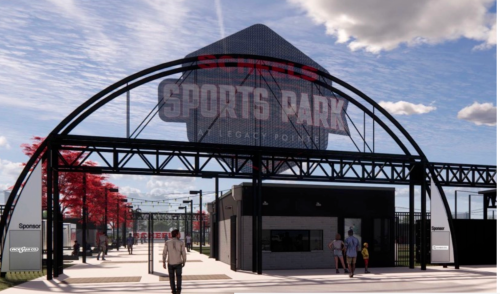 Entrance to a modern sports park with a large sign, surrounded by trees and a clear blue sky.