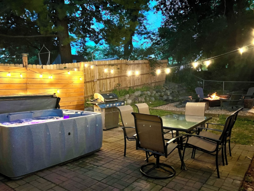 A cozy backyard scene with a hot tub, dining table, grill, and string lights, surrounded by trees and a fire pit.