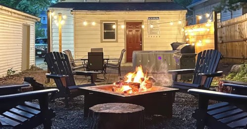 Cozy backyard scene with a fire pit, wooden chairs, string lights, and a grill, creating a warm, inviting atmosphere.