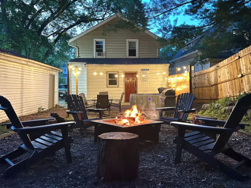 Cozy backyard scene with a fire pit surrounded by chairs, illuminated by string lights, and a house in the background.