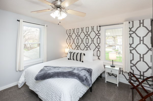 Cozy bedroom with a queen bed, decorative wall, two windows, and a ceiling fan. Soft gray blanket on the bed.