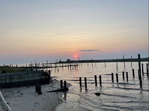 A serene sunset over calm waters, with wooden posts and a distant shoreline silhouetted against a colorful sky.