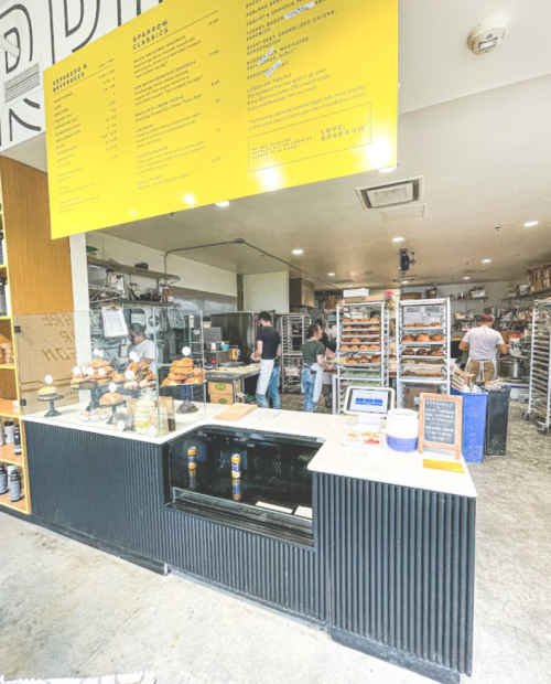 A bakery counter with a menu overhead, showcasing baked goods and staff working in a busy kitchen behind.