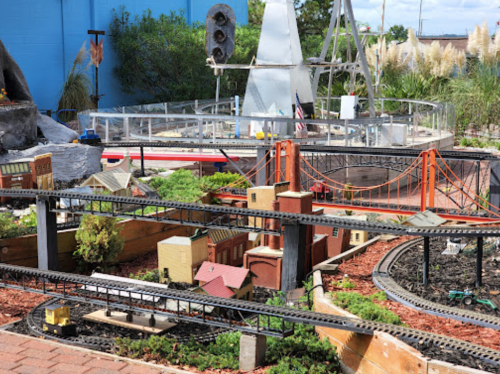 A miniature train setup featuring buildings, bridges, and tracks in a landscaped outdoor area.