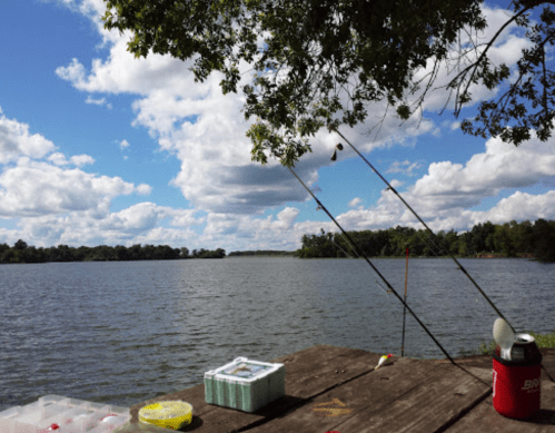 A serene lake view with fishing rods, a tackle box, and a cooler on a wooden dock under a cloudy sky.
